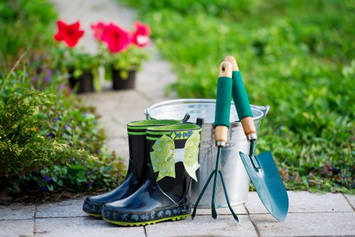 A close-up view of a garden pathway showing a pair of black rubber wellington boots with green patterned socks hanging inside, a small metallic hand trowel, a gardening fork, and a mini spade with teal handles leaning against a white plastic bucket, set against a background of lush green grass, flower beds with vibrant pink and purple blossoms, and a blurred garden scene. The area appears well-maintained with a paved surface, and the scene suggests outdoor gardening activities typical of a residential garden in Muswell Hill, with rich natural tones and a neat layout suitable for landscaping and garden care services. The lighting indicates a bright, possibly sunny day, highlighting the freshness of the garden environment.